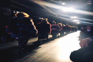 Eine Gruppe von Menschen geht über einen hell beleuchteten Laufsteg bei einer Modenschau, mit Zuschauern auf beiden Seiten.