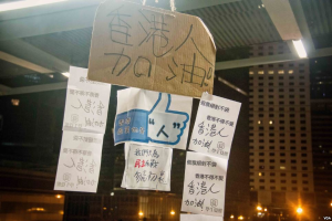 Eine Gruppe von Menschen steht vor einem Gebäude in der Nacht, beleuchtet von Stadtlichtern, mit mehreren hängenden Schildern, die wahrscheinlich gegen eine Regierungsentscheidung zur Social-Media-Sperre protestieren.