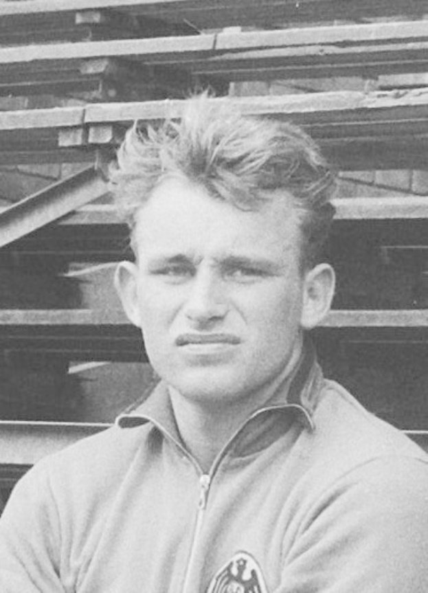 Schwarzes und weißes Foto eines Mannes in einer Jacke vor einem Stapel Holzplanken, der als legendärer deutscher Fussballer gilt.