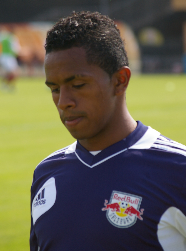 Junger Mann in einem blauen T-Shirt auf einem Fußballfeld, der als Flügelspieler mit kurzen dunklen Haaren und einem entschlossenen Gesichtsausdruck erscheint.