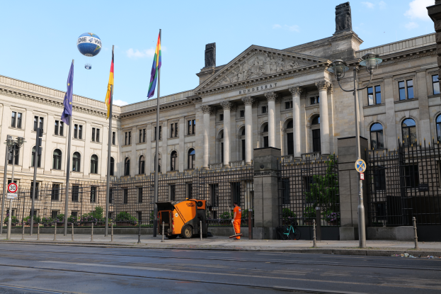 Das Reichstagsgebäude in Berlin, Deutschland, ein großes Gebäude mit zahlreichen Fenstern, umgeben von Straßenlaternen, Fahrzeugen, Menschen und Bäumen unter einem bewölkten Himmel.