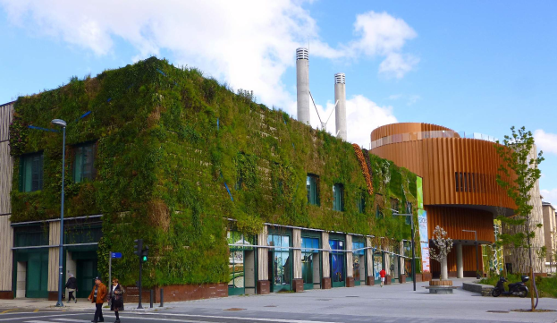 Ein großes Gebäude mit einer grünen Wand an seiner Seite, umgeben von Straßenmasten, Straßenlaternen, Verkehrspfosten, Verkehrssignalen, Kraftfahrzeugen, Fußgängern, Bäumen und einem bewölkten Himmel, Teil des Green Building Project.