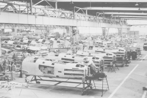 Ein Schwarz-Weiß-Foto einer belebten Fabrikhalle voller Flugzeuge und Arbeitern auf dem Boden, mit Lampen an der Decke.