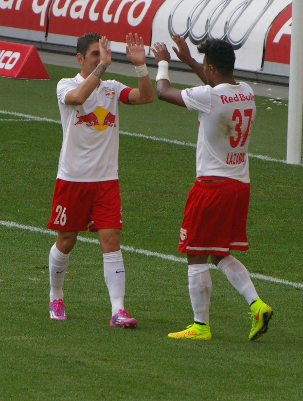 Zwei Fußballspieler stehen auf einem grünen Feld und klatschen sich ab, im Hintergrund eine Stange und eine Tafel mit Text.