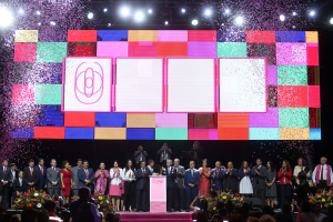 Gruppe von Menschen auf einer Bühne mit einem Podium, Blumengestecken, Kameras und einem großen Bildschirm, der Lichter für die Eröffnungszeremonie des 2018 China International Film Festivals anzeigt.