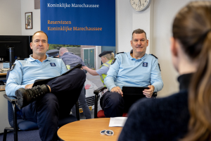 Zwei Polizeibeamte sitzen an einem Tisch mit einer Frau, umgeben von Dokumenten und Gegenständen, mit einem Wandbanner, einer Uhr und Fotorahmen im Hintergrund und einem zweiten Tisch mit Computerausrüstung links.