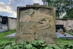 Ein steinernes Denkmal mit einer Skulptur einer Frau und eines Kindes, umgeben von Pflanzen am Boden, mit einer Wand im Hintergrund und Bäumen und Wolken am Himmel.