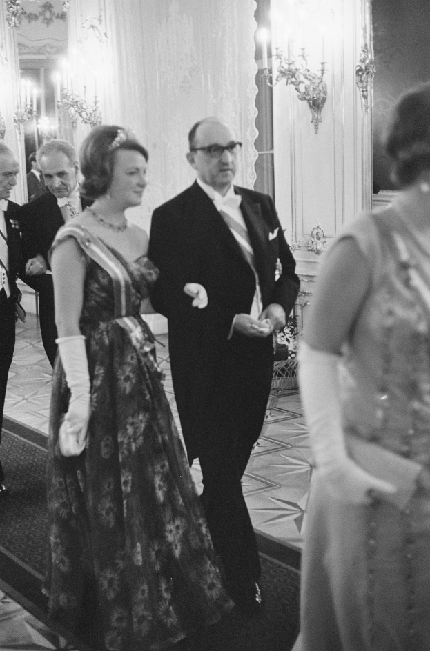 Ein Mann in einem schwarzen Blazer, weißem Hemd und schwarzen Hosen sowie eine Frau in einem eleganten Kleid gehen einen von Wandlampen beleuchteten Flur entlang, umgeben von einer Gruppe stehender Menschen.