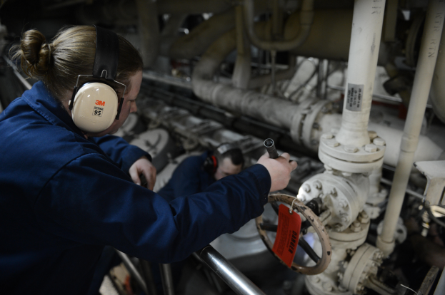 Zwei Frauen in blauen Uniformen bedienen Maschinen in einer Öl- und Gasanlage, eine hält ein Objekt und trägt Kopfhörer, mit Rohren und Equipment im Hintergrund.