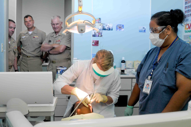 Eine Gruppe von Menschen in einer Zahnarztpraxis, mit einer Person in einem weißen Kleid, die ein Objekt hält, einer anderen mit Brille, Maske und einem Ausweis, sowie Plakate an der Wand im Hintergrund.