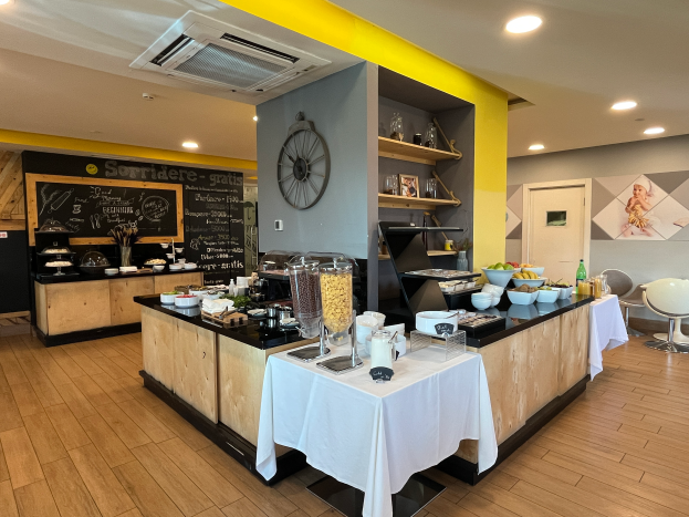 Ein Restaurantinnenraum mit Tischen und Stühlen, Speisen, einer Tafel mit Text, einer Wanduhr, Deckenleuchten, einer Klimaanlage und einer Tür im Hintergrund, befindet sich im Hotel de Ville in Paris.