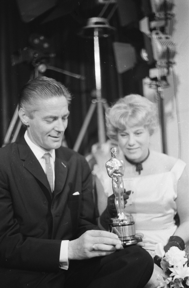 Ein Mann in einem schwarzen Blazer, wei├čem Hemd und Krawatte hölt eine Oscar-Statue, waehrend er neben einer Frau in Abendgarderobe sitzt, die einen Blumenstrau├č hölt, beide in einem Schwarz-Wei├č-Bild.