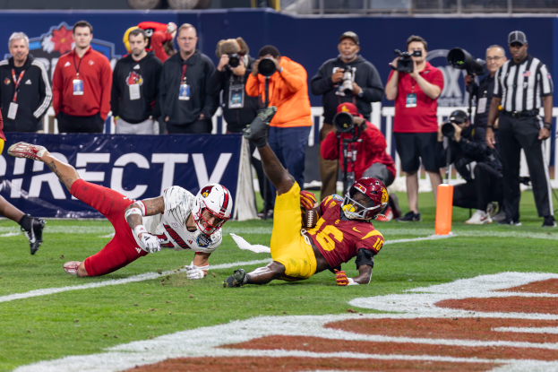 Ein Fußballspieler taucht in die Endzone für einen Touchdown ab, mit einer Gruppe von Menschen am Boden, einige halten Kameras, und ein Banner mit Text im Vordergrund, während Geländer, Pfosten und andere Gegenstände im Hintergrund sichtbar sind.
