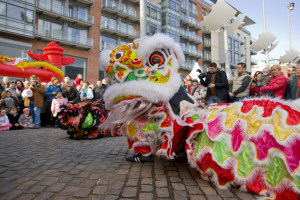 Vibrierendes chinesisches Neujahrsfest in Amsterdam mit einer Löwen-Tanz-Performance im Vordergrund und einer Menschenmenge drumherum, einige halten Kameras, vor einem Hintergrund aus Gebäuden, Laternenmasten und einem klaren blauen Himmel.