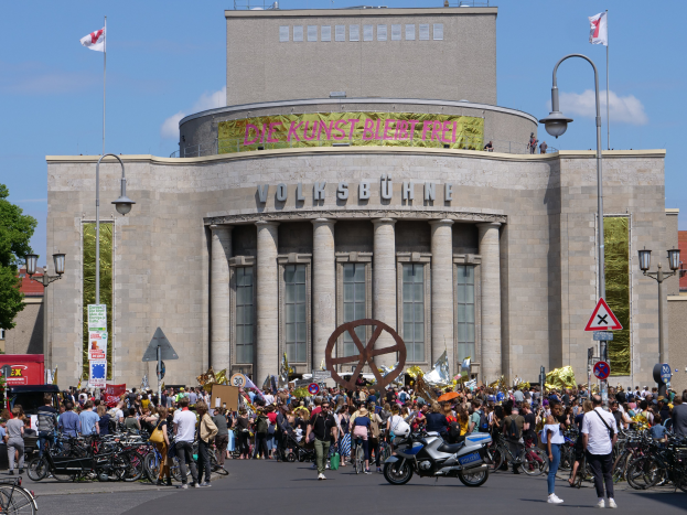 Eine große Gruppe von Menschen versammelt sich vor einem Gebäude mit Säulen und Text, umgeben von Straßenlaternen, Schildern, Motorrädern, Fahrzeugen, Bäumen und einem bewölkten Himmel während einer Demonstration in Berlin, Deutschland.