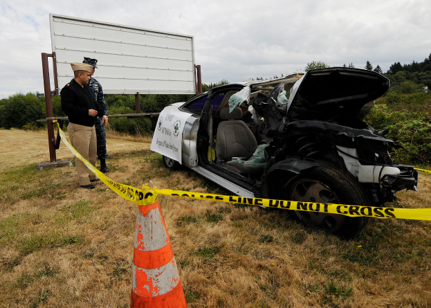 Ein Polizeibeamter in Uniform steht neben einem schwer beschädigten Auto am Straßenrand, mit einem Verkehrskegel und einem Band an einem Pfahl in der Nähe und Bäumen und einem bewölkten Himmel im Hintergrund.