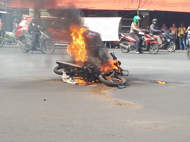 Motorrad in Flammen auf der Straße mit einem Auto und einem Van im Hintergrund, umgeben von Menschen, einige mit Helmen, eine Fahne mit Text und ein Zelt.
