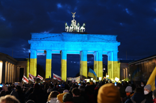 Eine Menschenmenge steht vor dem Brandenburger Tor in Berlin und hält Fahnen, Schilder und ein Banner mit Text unter einem bewölkten Himmel.