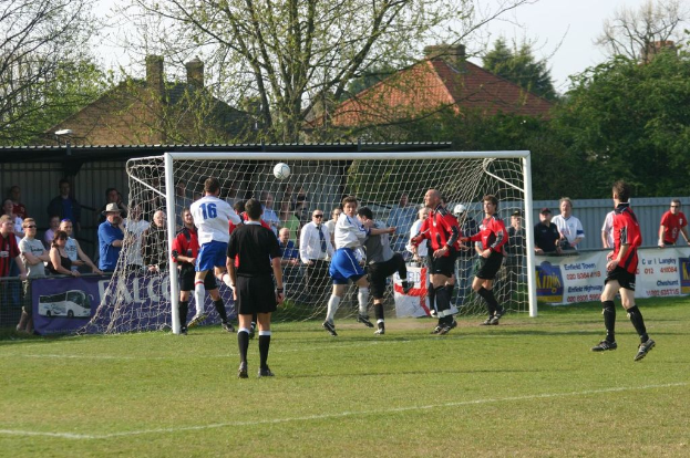 Spieler sind in einem Fussballspiel auf einem Feld mit einem Tornetz beteiligt, während Zuschauer dahinter stehen, mit Bäumen und Häusern im Hintergrund.