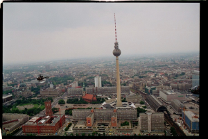 Eine Luftaufnahme von Berlin, Deutschland, mit dem Fernsehturm in der Mitte, umgeben von Gebäuden, Bäumen, Straßen, Fahrzeugen und einem Hubschrauber unter einem klaren blauen Himmel.