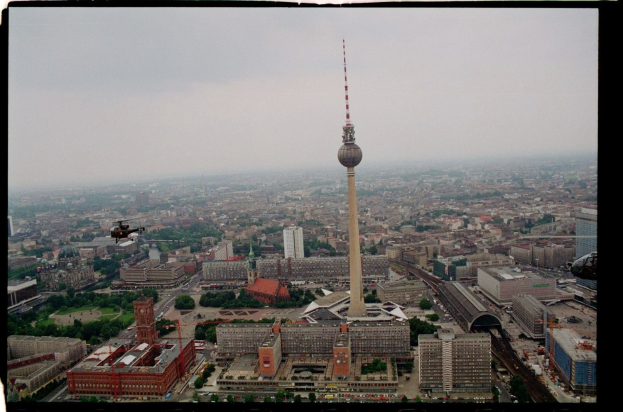 Eine Luftaufnahme von Berlin, Deutschland, mit dem Fernsehturm in der Mitte, umgeben von Gebäuden, Bäumen, Straßen, Fahrzeugen und einem Hubschrauber unter einem klaren blauen Himmel.