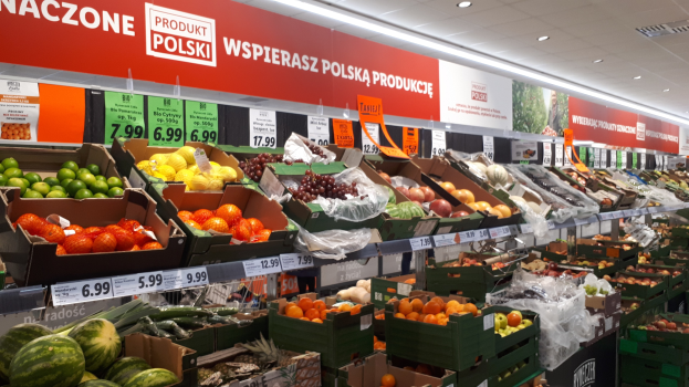 Ladeninneres mit verschiedenen Früchten und Gemüsen in Kartonschachteln, jede mit Preisschild, Wandtafeln mit Text und Bildern und Deckenbeleuchtung.