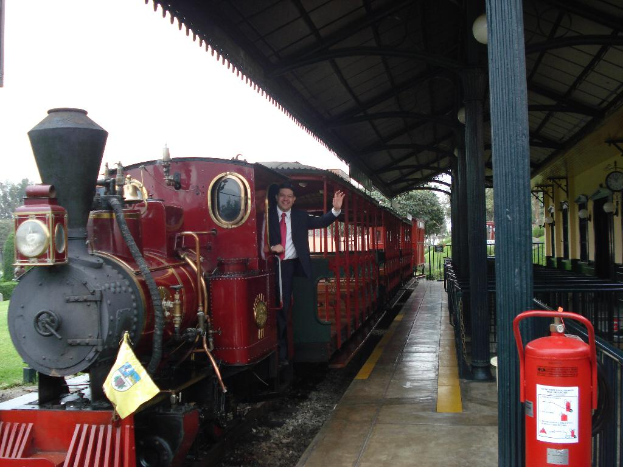 Ein roter Zug mit einem Mann in einem schwarzen Anzug an der Tür, der lächelt und posiert, mit einem Bahnhofsgebäude auf der rechten Seite und einem roten Feuerlöscher im Vordergrund.