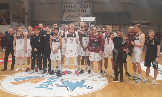 Eine Gruppe von Männern und Kindern auf einem Basketballfeld mit Schildern, die "Hala Osir Starga" lesen, sitzende Zuschauer und Basketballausrüstung im Hintergrund.