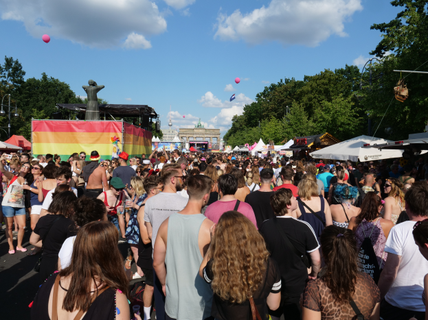 Eine große Menschenmenge geht eine Straße mit Zelten, Bäumen, Pfählen, Lichtern und einer Statue entlang, im Hintergrund Gebäude und ein Himmel mit Wolken und Ballons bei der Berlin Pride.
