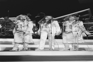 Drei Frauen singen in Mikrofone auf der Bühne mit Musikern, die hinter ihnen spielen.