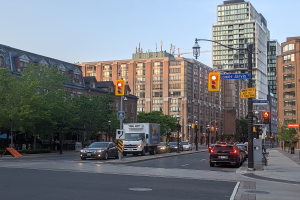 Eine belebte Stadtstraße mit verschiedenen Fahrzeugen einschließlich einem Lastwagen, einem Auto und einem SUV, umgeben von Laternenmasten, Ampeln, Texttafeln, Bäumen, Gebäuden mit Fenstern und einem Himmel im Hintergrund.