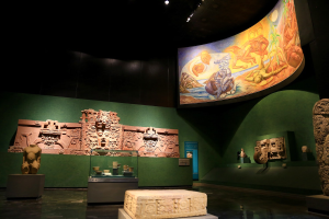 Ein Museumsraum mit verschiedenen Artefakten, darunter Skulpturen, Tischgegenstände, eine Texttafel, eine Wandmalerei und Deckenleuchten im Anthropologischen Museum von Mexiko-Stadt.