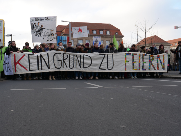 Demonstranten halten ein Transparent mit der Aufschrift "Kein Grund zu Feiern" gegen deutsche Sparmaßnahmen, mit Gebäuden, Bäumen und einem klaren Himmel im Hintergrund.
