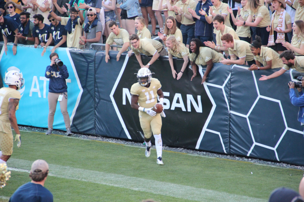 Ein Fußballspieler läuft mit dem Ball auf einem grünen Rasenfeld vor einer Menge, einige halten Kameras und tragen Helme, mit einem Banner im Hintergrund.