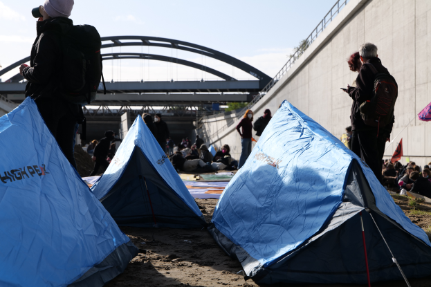 Eine Gruppe von Menschen sitzt auf einem sandigen Strand in der Nähe von Zelten, mit einer Wand auf der rechten Seite und einer Brücke im Hintergrund, die Gegenstände halten und Taschen tragen während einer Klimawandel-Demonstration unter einem bewölkten Himmel.