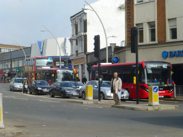 Ein roter Doppeldeckerbus fährt auf einer von hohen Gebäuden gesäumten Stadtstraße, umgeben von Fußgängern, einem Fahrradfahrer, Ampeln, Laternen und Schildern unter einem bewölkten Himmel.