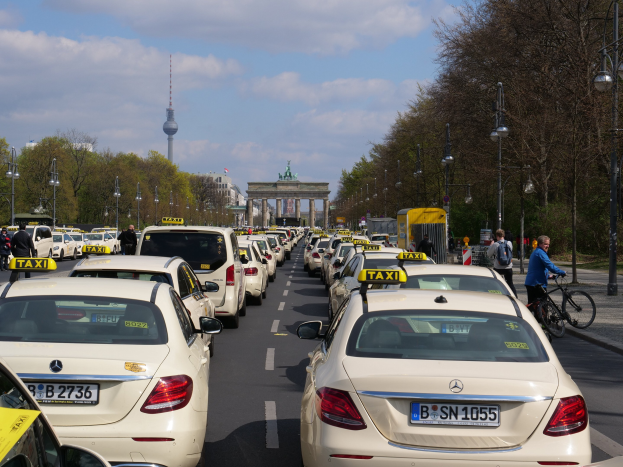 Eine lange Reihe von Taxis, die entlang einer belebten Straße in Berlin, Deutschland, geparkt sind, mit Fahrzeugen, Radfahrern und Fußgängern, flankiert von Bäumen und Laternenpfählen und Gebäuden, einem Bogen und einem Turm im Hintergrund unter einem bewölkten Himmel.