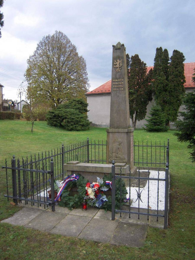 Ein Denkmal auf einer grünen Wiese mit einem Kranz, umgeben von einem Metallzaun, erinnert an Holocaust-Opfer in Vostiu, Polen, mit Bäumen, Gebäuden und einem klaren blauen Himmel im Hintergrund.