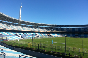 Ein großes Stadion mit einem Fußballfeld, das von einem Zaun umgeben ist, mit einem Turm im Hintergrund unter einem klaren blauen Himmel.