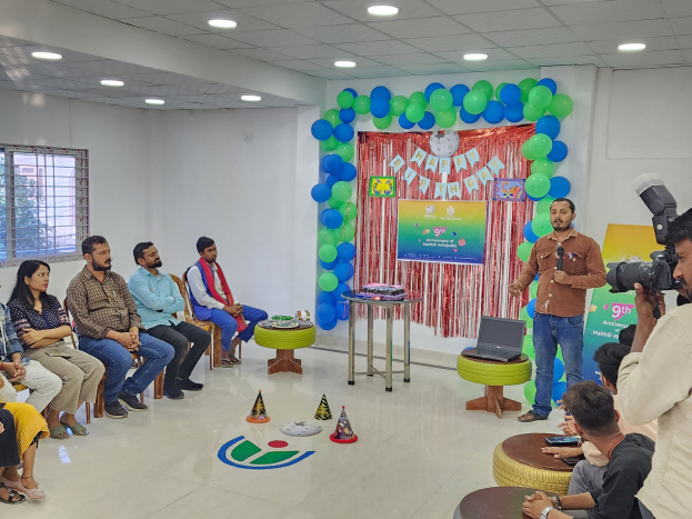 Eine Gruppe von Menschen, die auf Stühlen sitzen und einer Menge gegenübersitzen, mit einer Person, die ein Mikrofon hält, einem Laptop auf einem Hocker, einem Tisch mit Gegenständen, einer Fahne, Ballons, einem Vorhang, einem Fenster und Deckenleuchten, was eine 9. Geburtstagsfeier vermuten lässt.