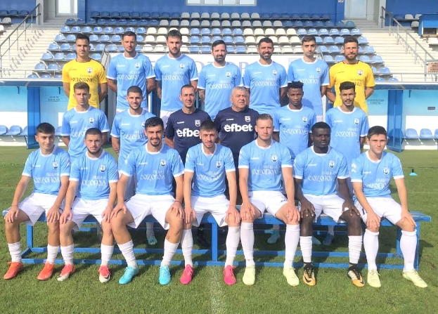 Gruppe von Männern auf einem Fuballfeld, einige sitzen auf Bänken und einige stehen auf dem Rasen, mit Tribünen und Gebäudestrukturen im Hintergrund.