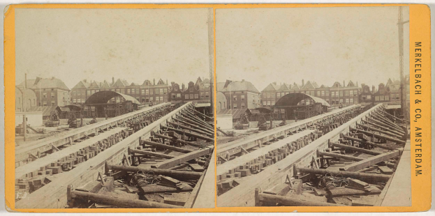 Ein altes Schwarz-Weiß-Foto einer Bahnschiene mit Gebäuden im Hintergrund, Text auf der rechten Seite mit 'Merkelbach & Co. Amsterdam' und verstreuten Holzobjekten im Vordergrund.