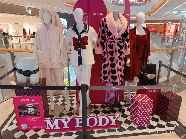 Eine Schaufensterdekoration mit Schaufensterpuppen auf einem schwarz-weißen Schachbrettboden, mit Menschen, Kleidung, Beschilderung und einer Tafel mit der Aufschrift "My Body", was auf einen Victoria's Secret-Laden hinweist.