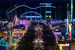 Eine große Menschenmenge steht um einen hell erleuchteten Karneval in der Nacht herum, umgeben von Vergnügungsgeräten, Pfählen, Flaggen und Namensschildern, mit einem dunklen Hintergrund, der eine festliche Atmosphäre schafft.