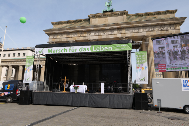 Eine Bühne vor dem Brandenburger Tor in Berlin, Deutschland, mit einem Tisch, einer Vase mit Blumen, einem Kreuzsymbol und anderen Gegenständen sowie Transparenten mit Text. Es gibt auch Lautsprecher und andere Gegenstände auf der Bühne und Fahrzeuge auf der Straße. Im Hintergrund sind Gebäude mit Fenstern, eine Statue auf einem Gebäude, ein Mast mit einer Fahne und ein bewölkter Himmel zu sehen.