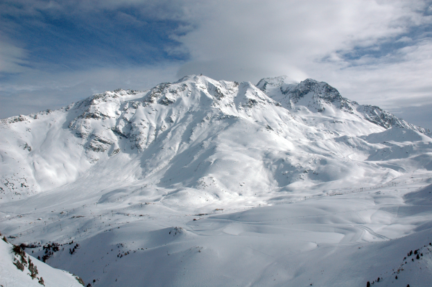 Ein schneebedeckter Berg mit ein paar Skifahrern, die hinunterfahren, umgeben von einer bewölkten Himmel und unberührtem Schnee.