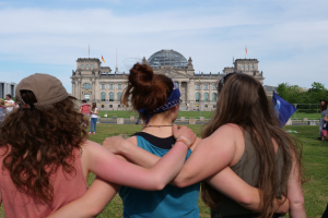 Drei Frauen in verschiedenen farbigen Kleidern, eine trägt eine Mütze, stehen vor dem Reichstagsgebäude in Berlin, Deutschland, mit Menschen, Fahnen, Bäumen, Wolken und Himmel im Hintergrund.