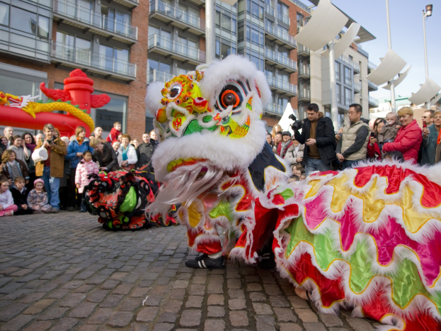 Ein lebendiges chinesisches Neujahrsfest in Amsterdam mit einer Löwen-Tanz-Show vor einer Zuschauermenge, darunter einige, die das Ereignis fotografieren, vor Buildings, Laternenmasten und einem klaren blauen Himmel.