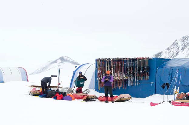 Drei Personen stehen auf einer verschneiten Landschaft mit verstreuten Taschen, Zelte mit Skiern dahinter und schneebedeckten Hügeln im Hintergrund unter einem klaren Himmel.