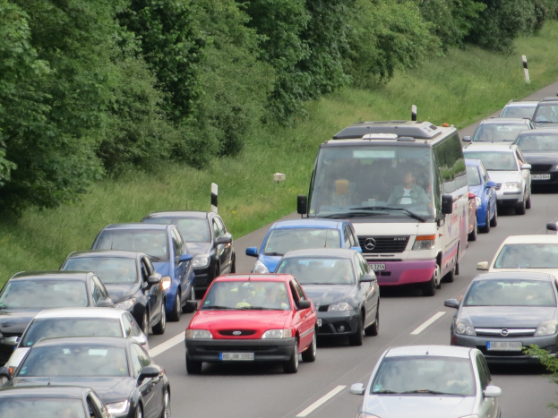 Ein Stau auf der Autobahn mit mehreren Autos und einem Van, mit Passagieren in den Fahrzeugen, vor dem Hintergrund von Bäumen und Gras.